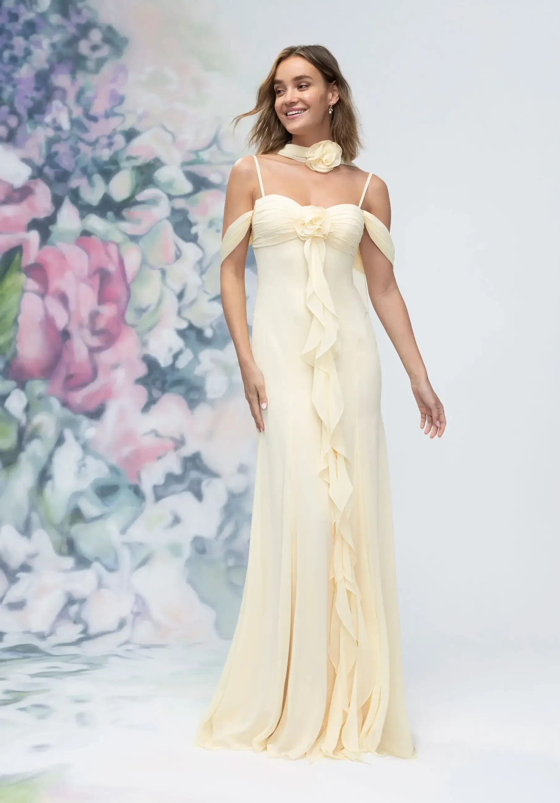 A model wearing a long, pale yellow dress with off-the-shoulder straps and ruffled details, standing in front of a floral background.