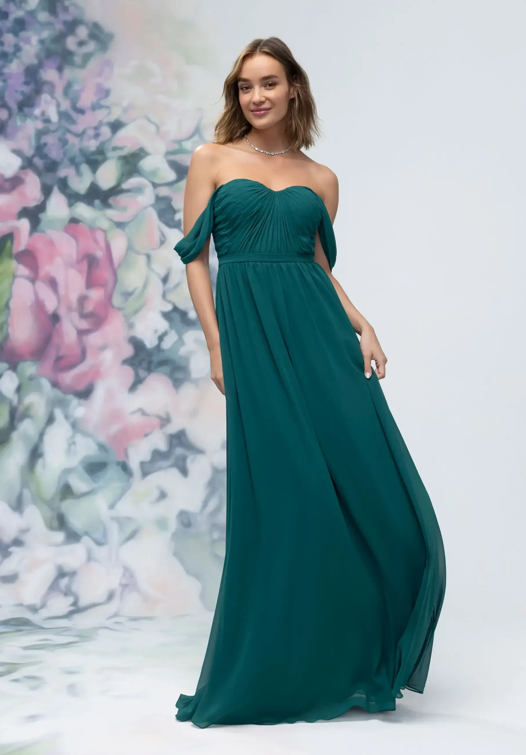 A woman is wearing a teal off-the-shoulder long dress, posing in front of a floral background.