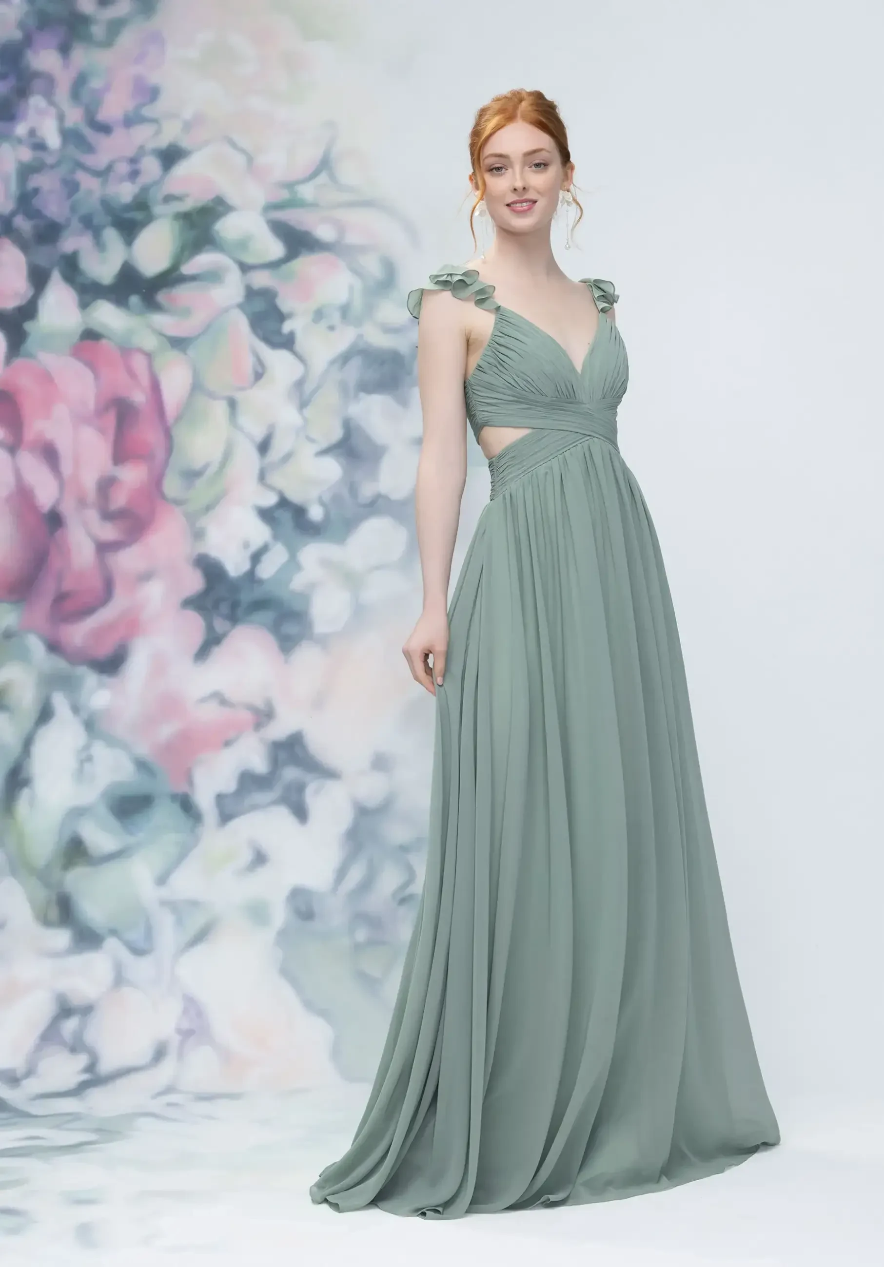 A model is wearing a long, flowy green dress with a V-neck and ruffled straps, standing against a floral background.