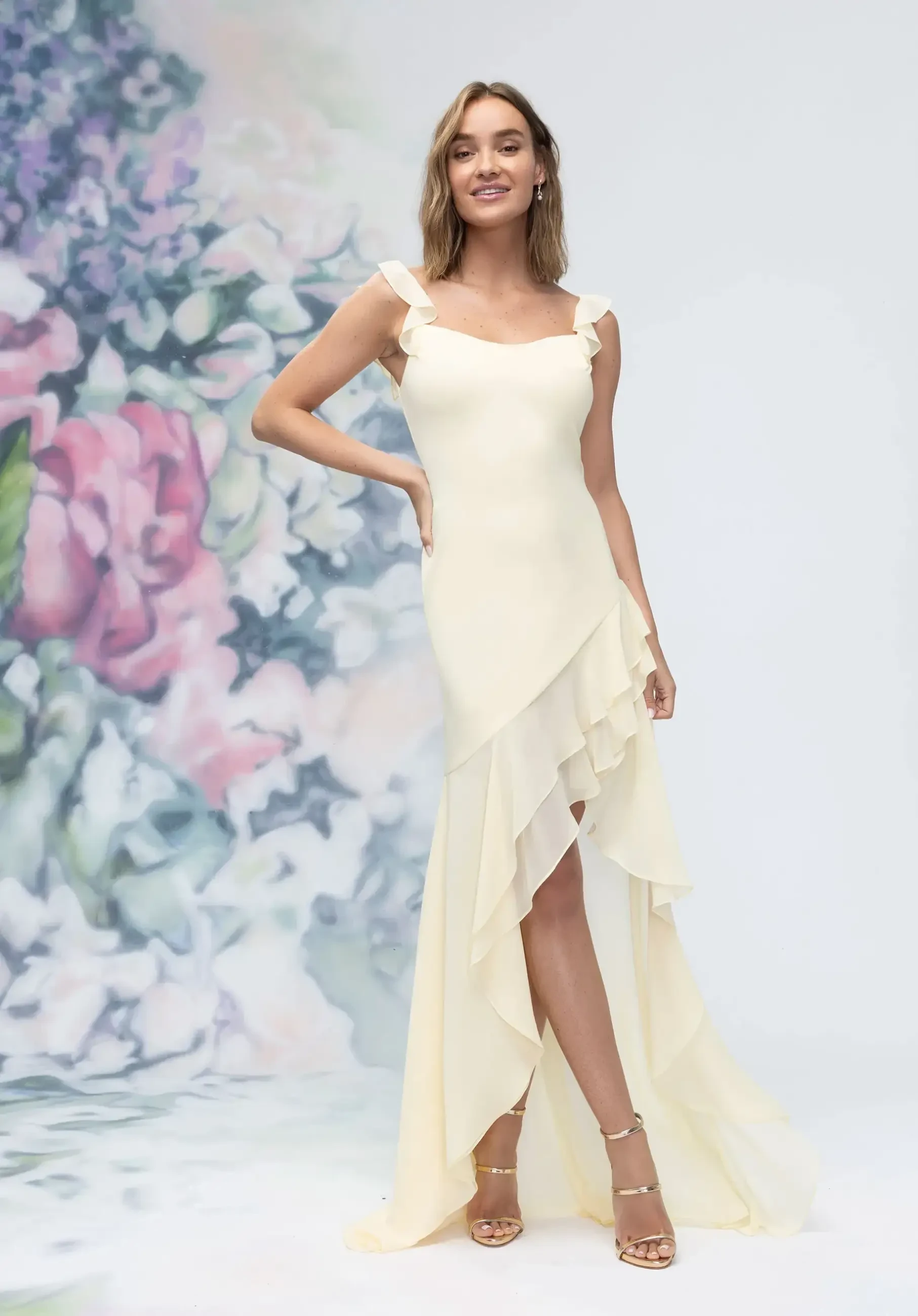 A woman is wearing a light yellow, elegant dress with ruffled straps and an asymmetrical hemline. She stands against a soft, floral background featuring pastel-colored flowers.