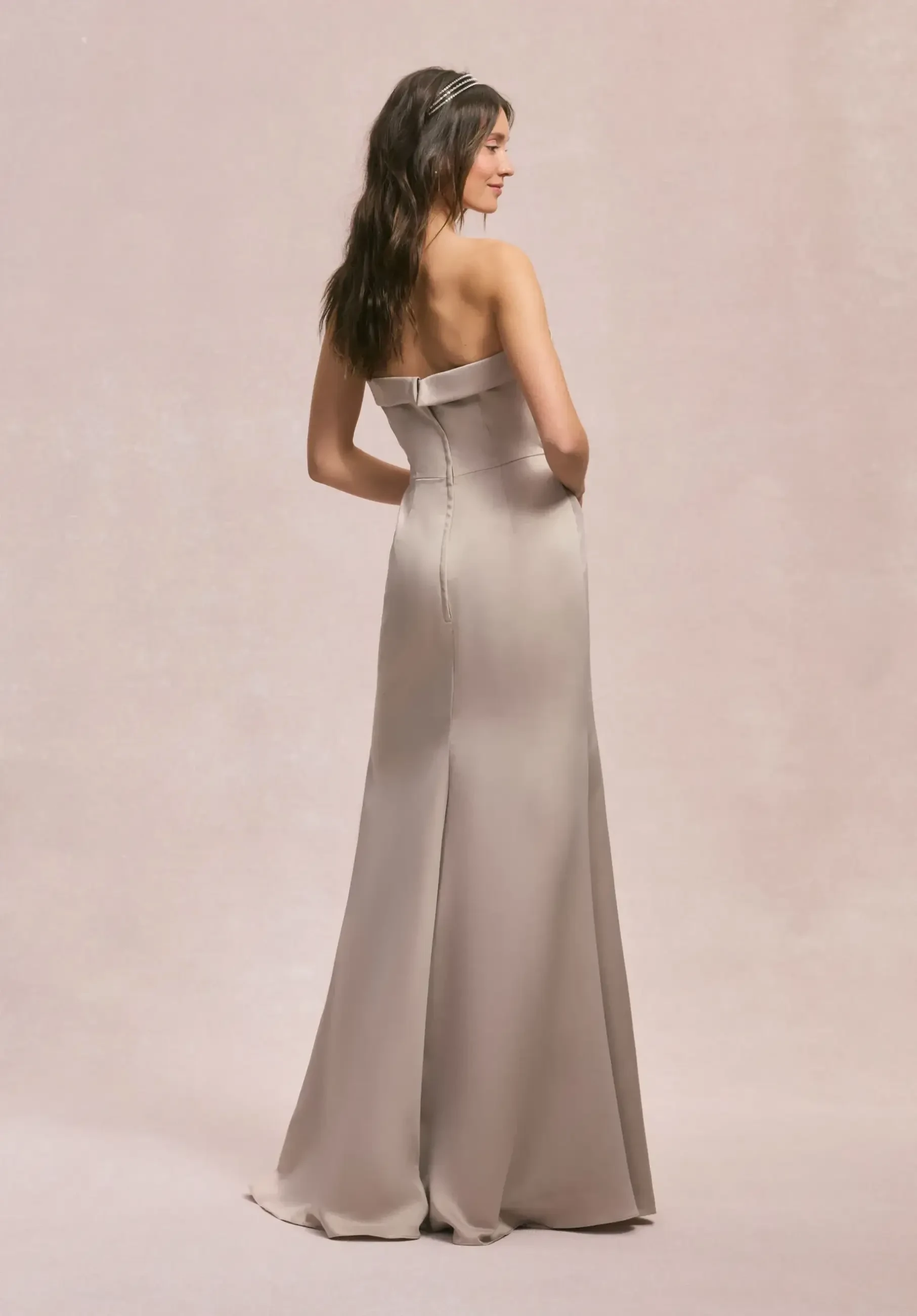 Model is wearing a strapless satin A-line bridesmaid dress with a sculptural bow detail at the waist, sleek clean lines, and pockets.