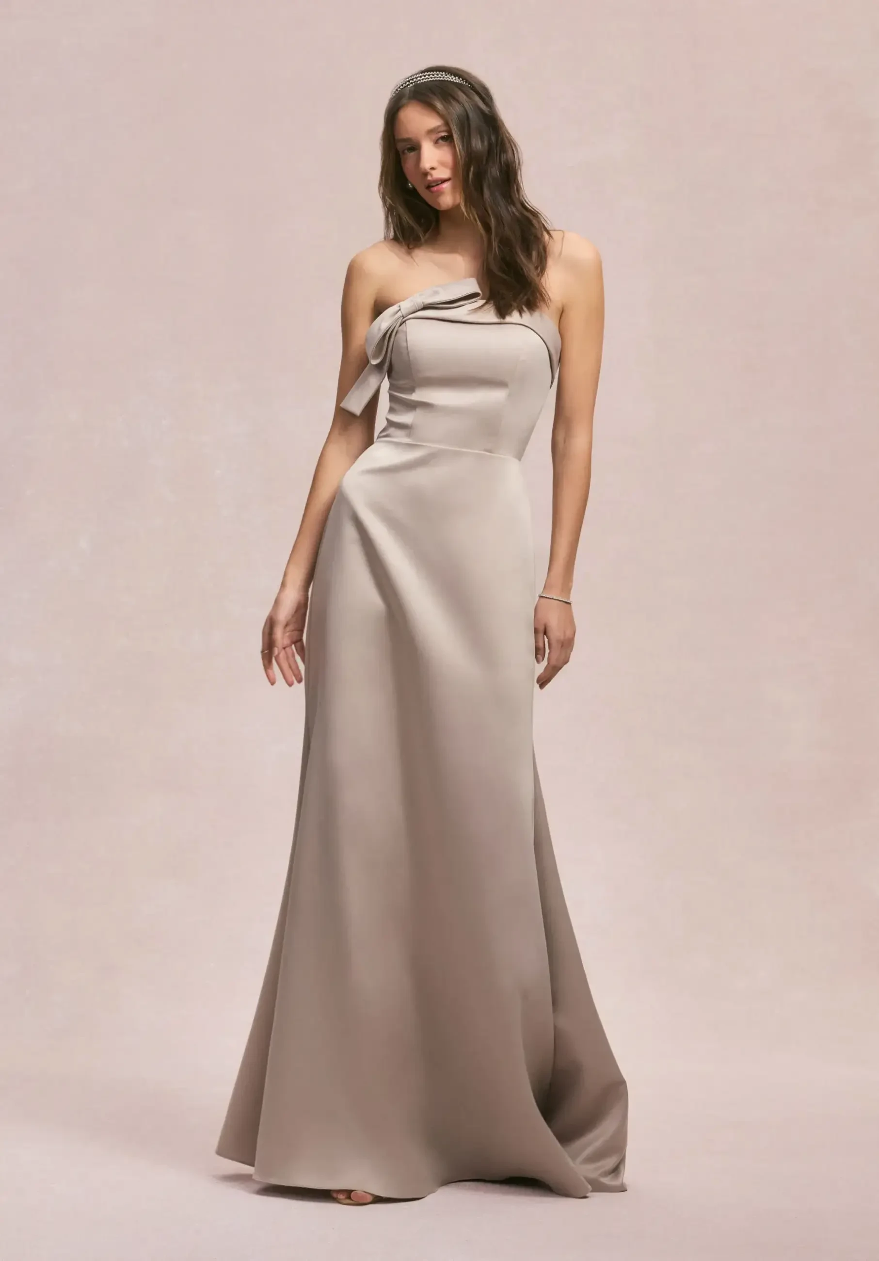 Model is wearing a strapless satin A-line bridesmaid dress with a sculptural bow detail at the waist, sleek clean lines, and pockets.