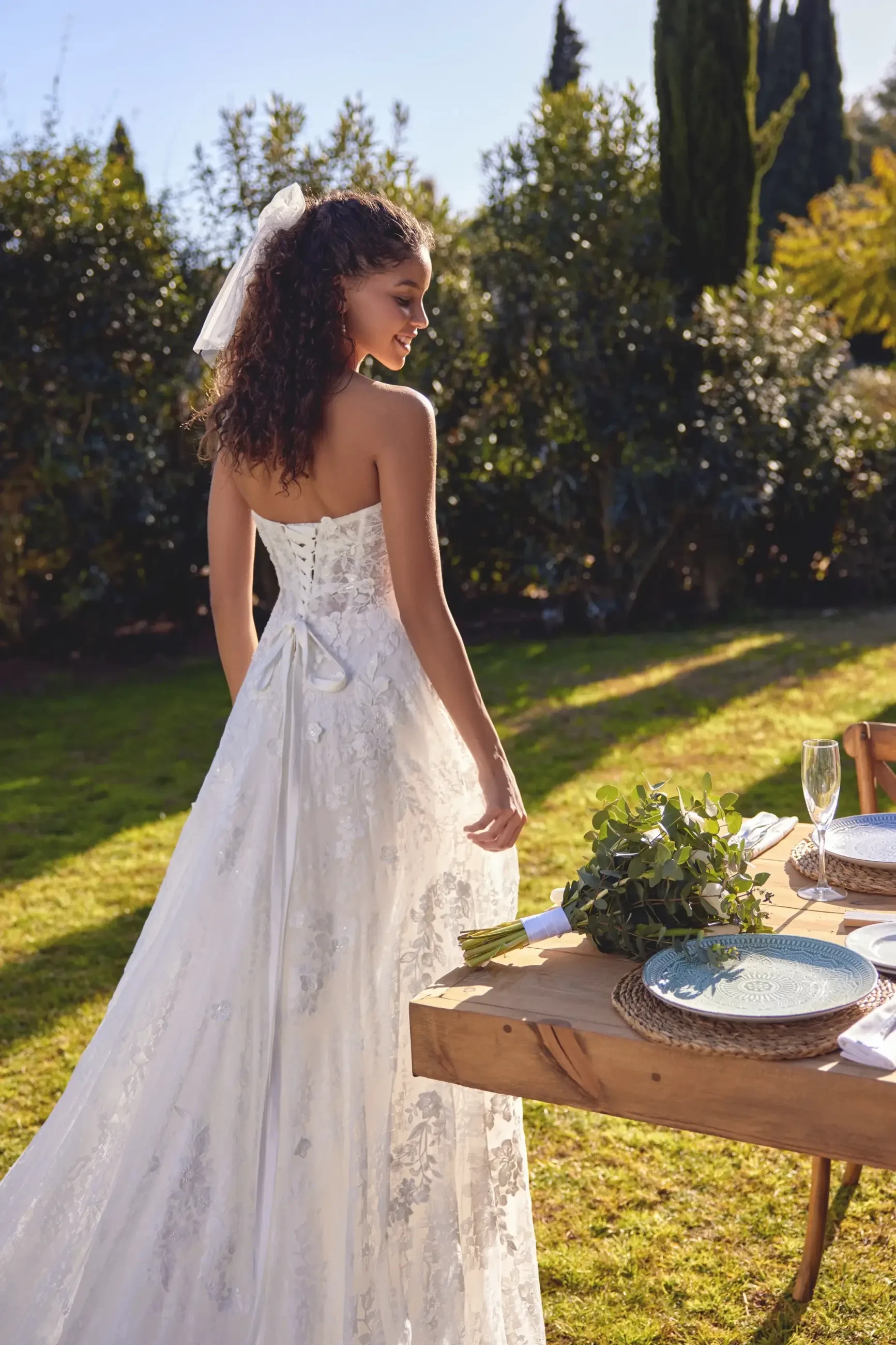 A person wearing a wedding dress stands in a garden, looking back. The scene includes an outdoor dining setup with a table, plates, and greenery.