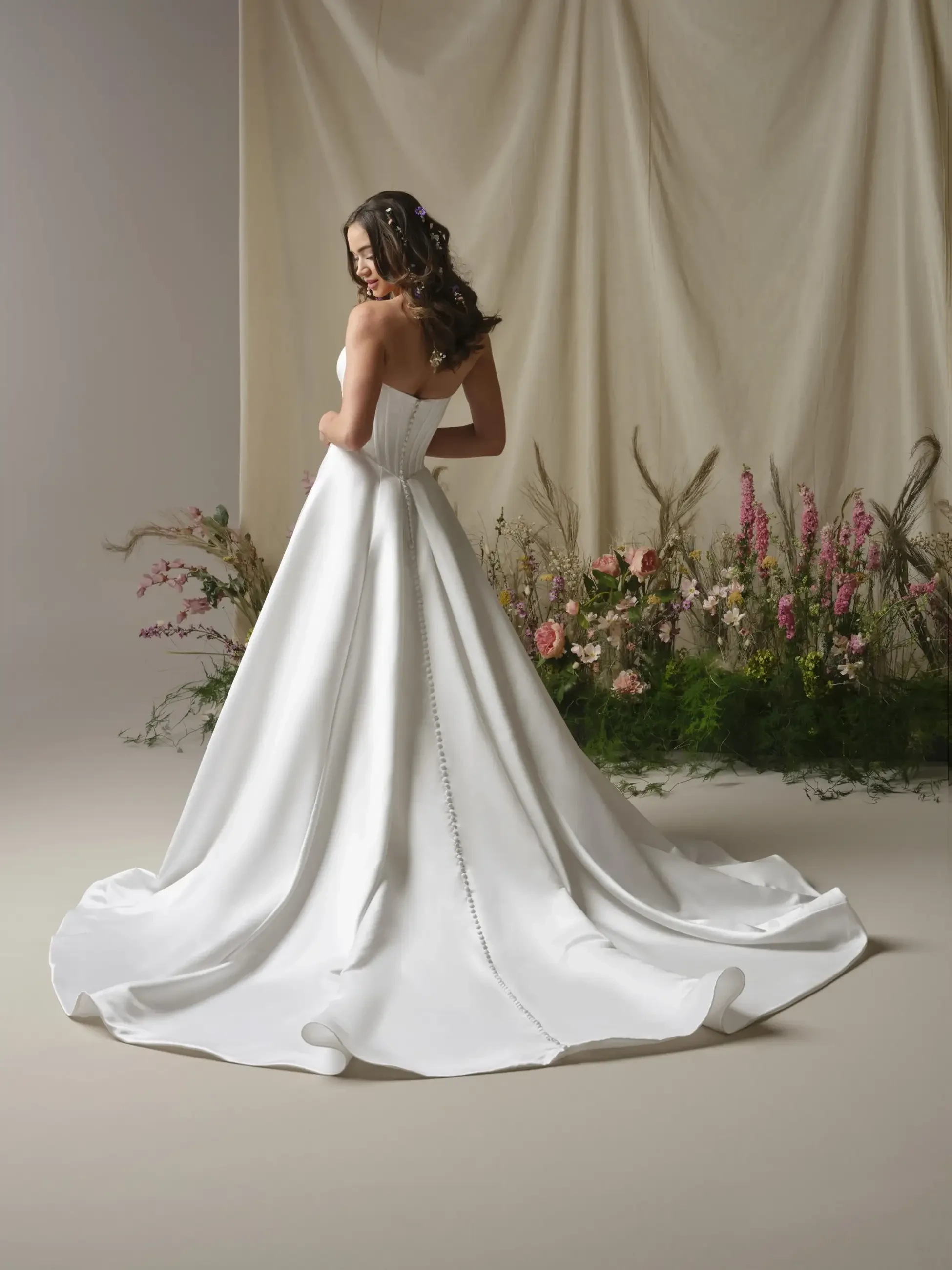 A model wearing a strapless white wedding gown, standing with her back to the camera, in front of a floral arrangement.