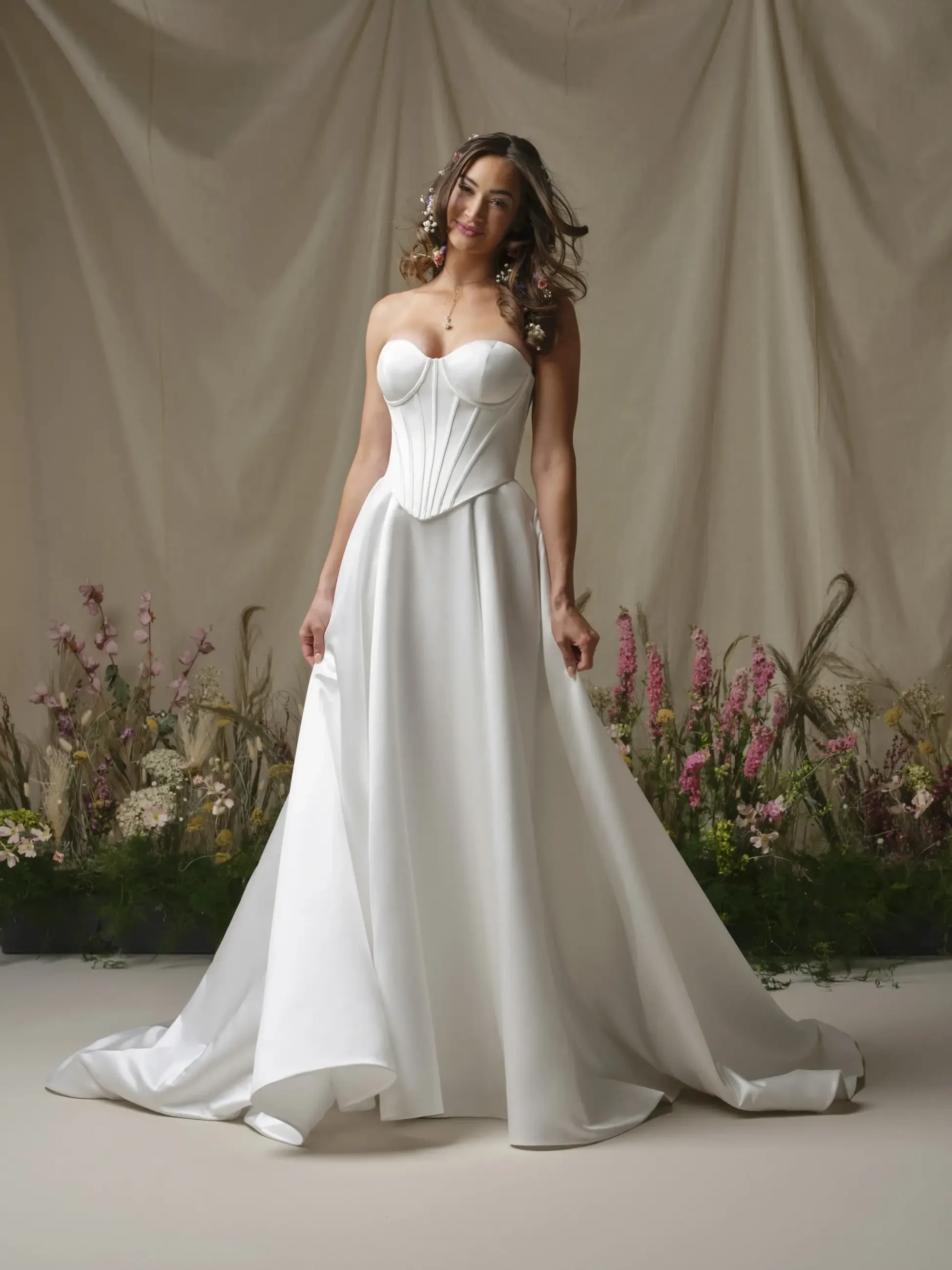 A woman in a strapless white gown standing on a neutral background with flowers and greenery around her.