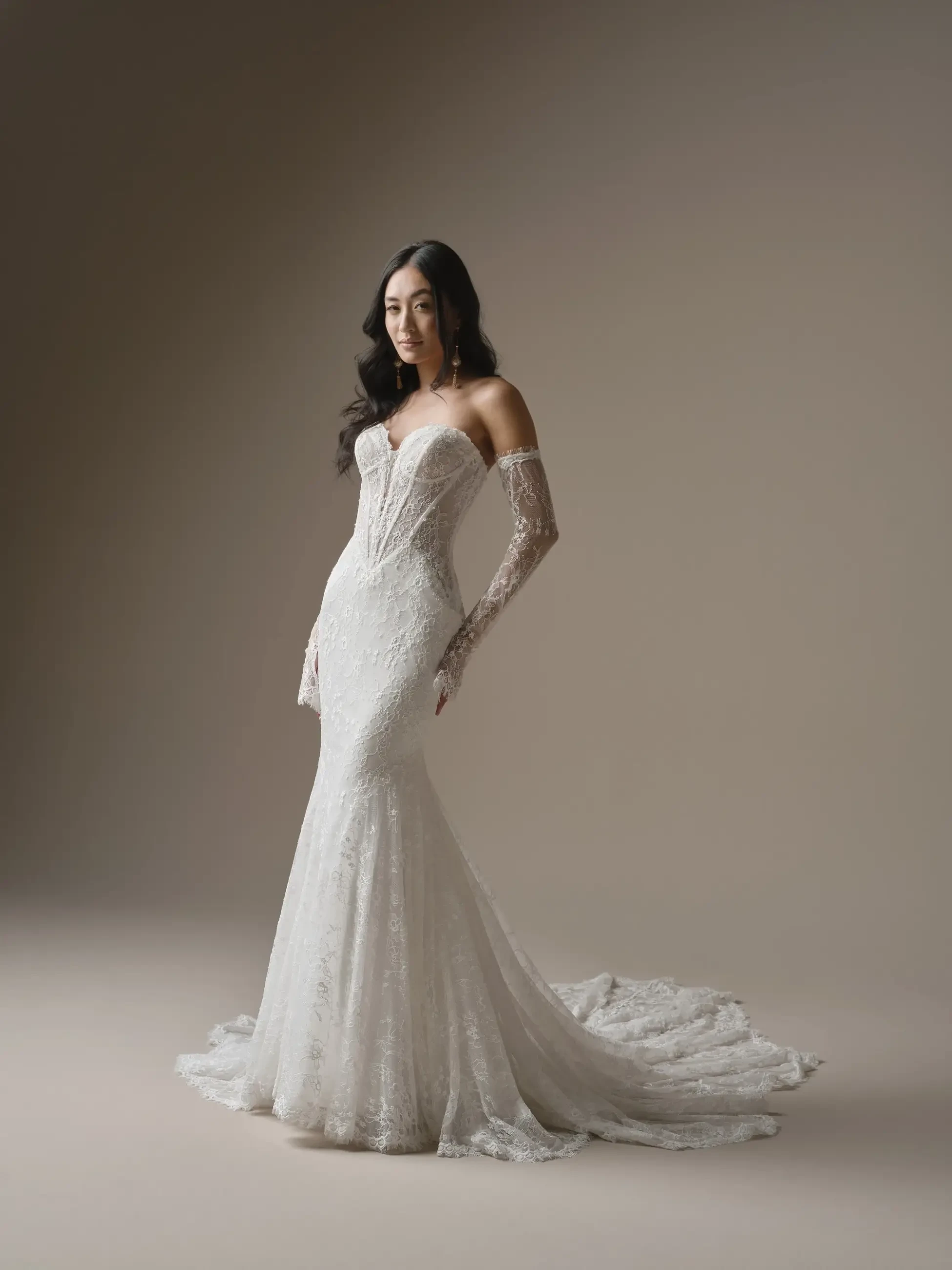 A woman in an elegant, off-the-shoulder lace wedding gown with long sleeves and a flowing train stands confidently against a neutral backdrop.