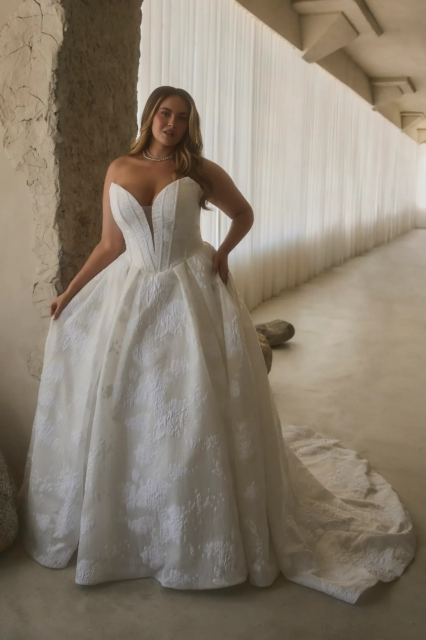 A woman wearing a strapless white wedding gown stands elegantly in a spacious area with soft lighting and draped fabric in the background.