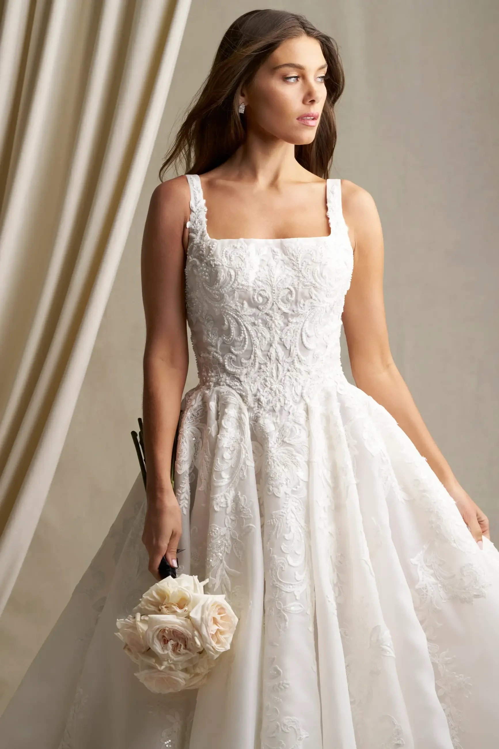 A woman in a detailed white lace wedding dress holds a bouquet of pale roses. She gazes thoughtfully to the side against a neutral, draped background.