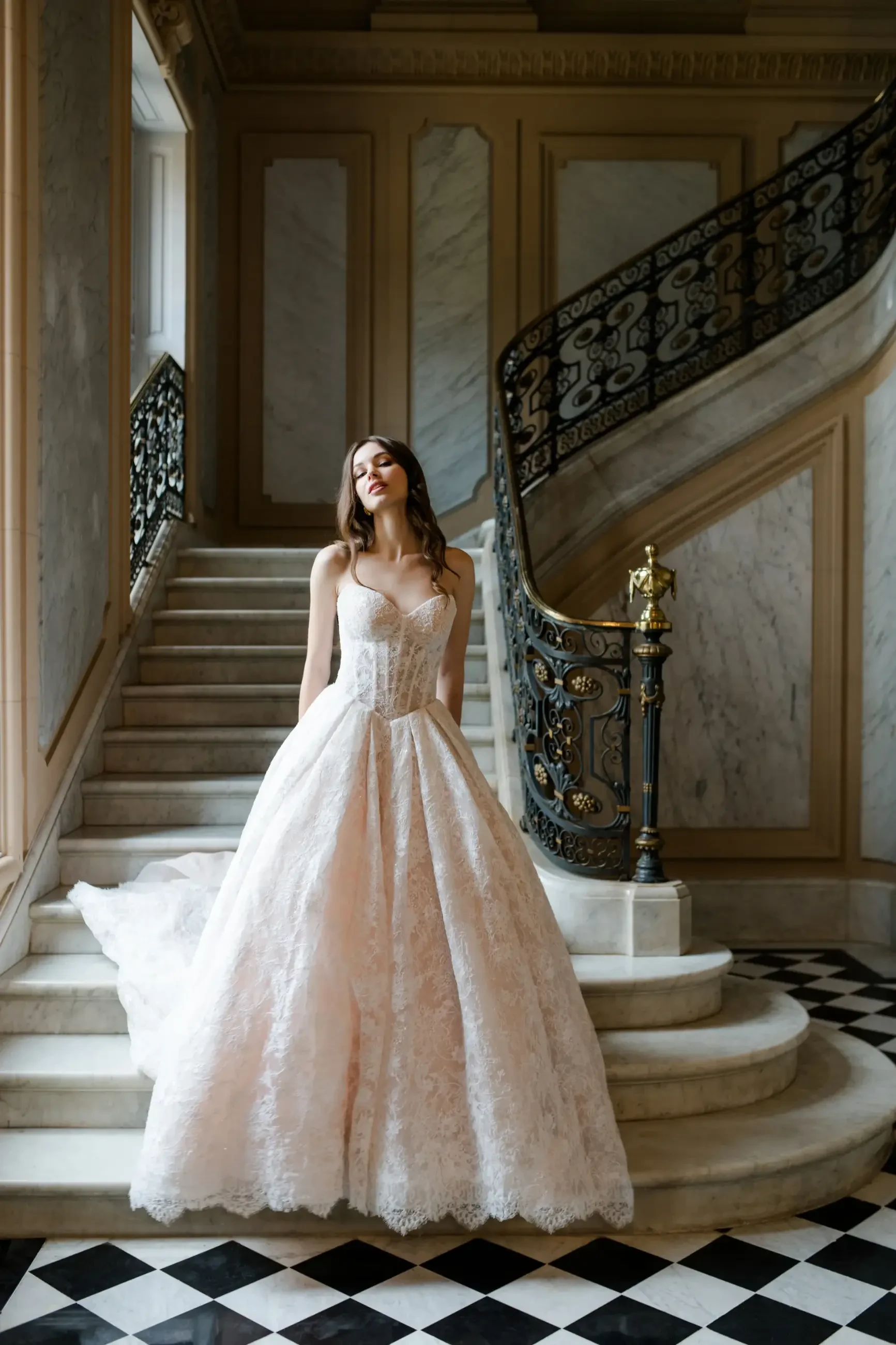 A woman in an elegant pink lace gown stands on marble stairs in a grand interior. The metal railing and checkered floor add a luxurious touch.