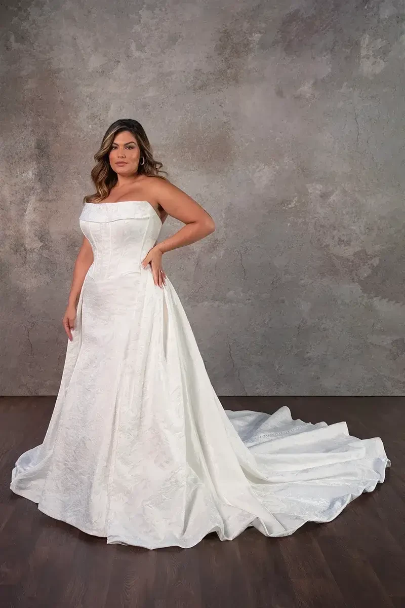 Model is wearing a plus size strapless fit and flare wedding dress with corset boning and a basque waist
