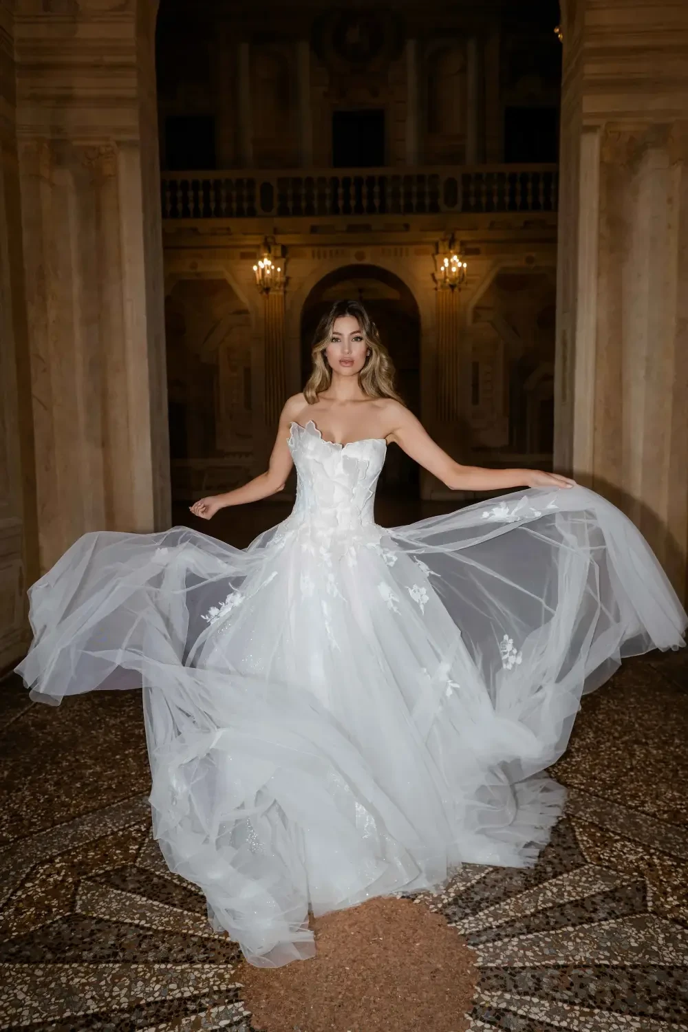 A woman in a wedding dress poses elegantly in an ornate, dimly lit interior, with flowing fabric creating a dramatic effect.