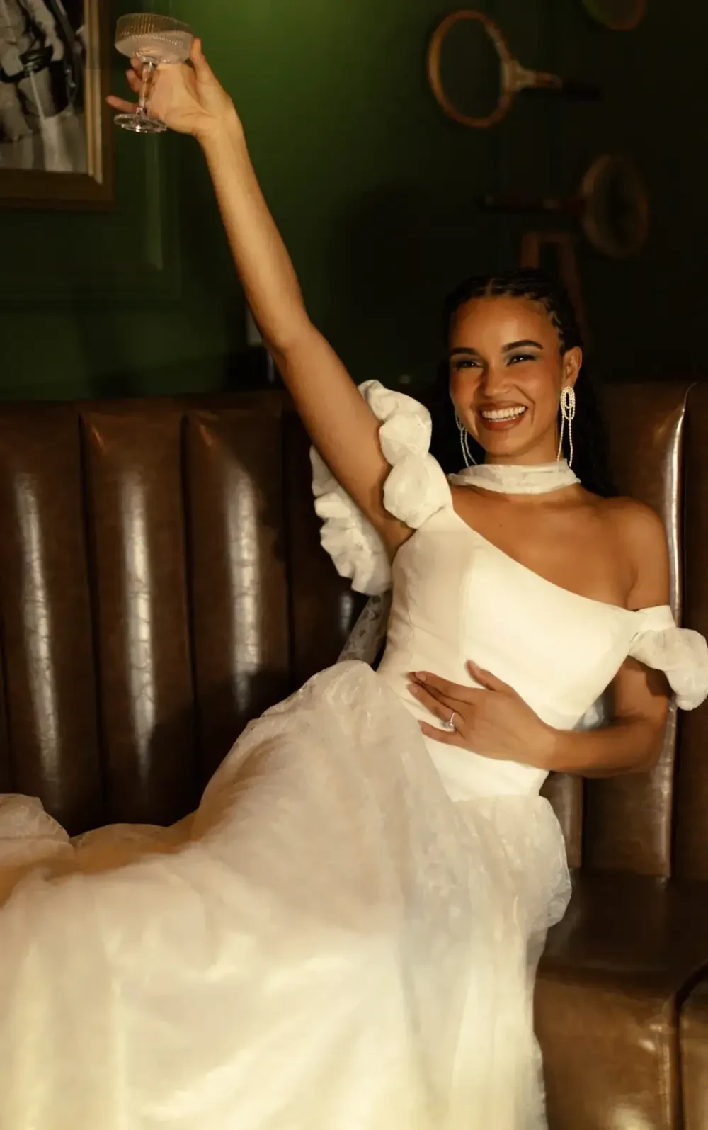 A woman in a white off-shoulder gown raising a glass, seated on a brown leather couch, smiling.