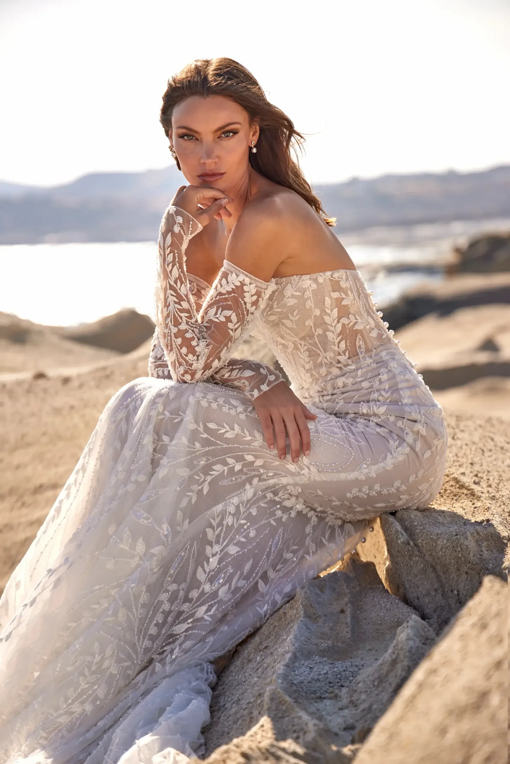 Woman in an elegant, lace-patterned, off-shoulder gown poses gracefully on rocky terrain by the sea. The serene backdrop and her contemplative expression create a tranquil, bridal atmosphere.