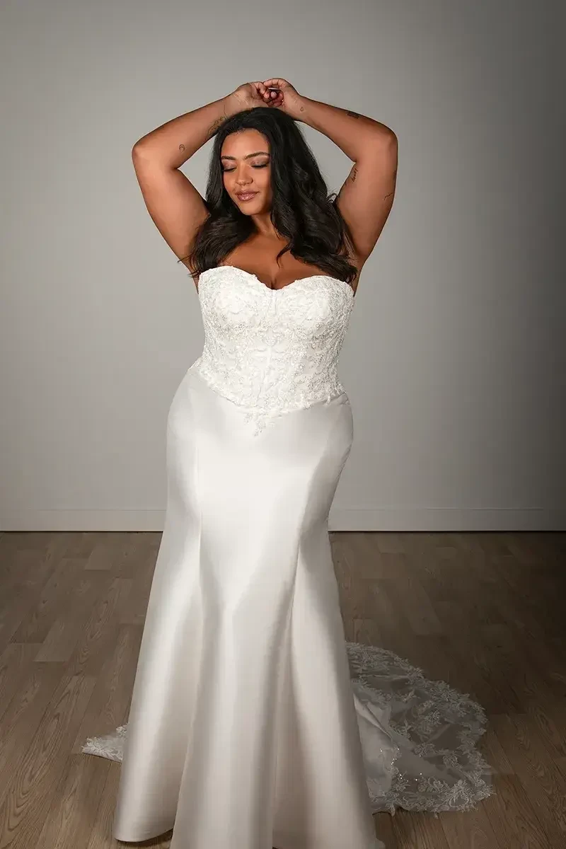 A woman wearing a strapless wedding gown, posing with her arms raised in a graceful manner against a neutral background.