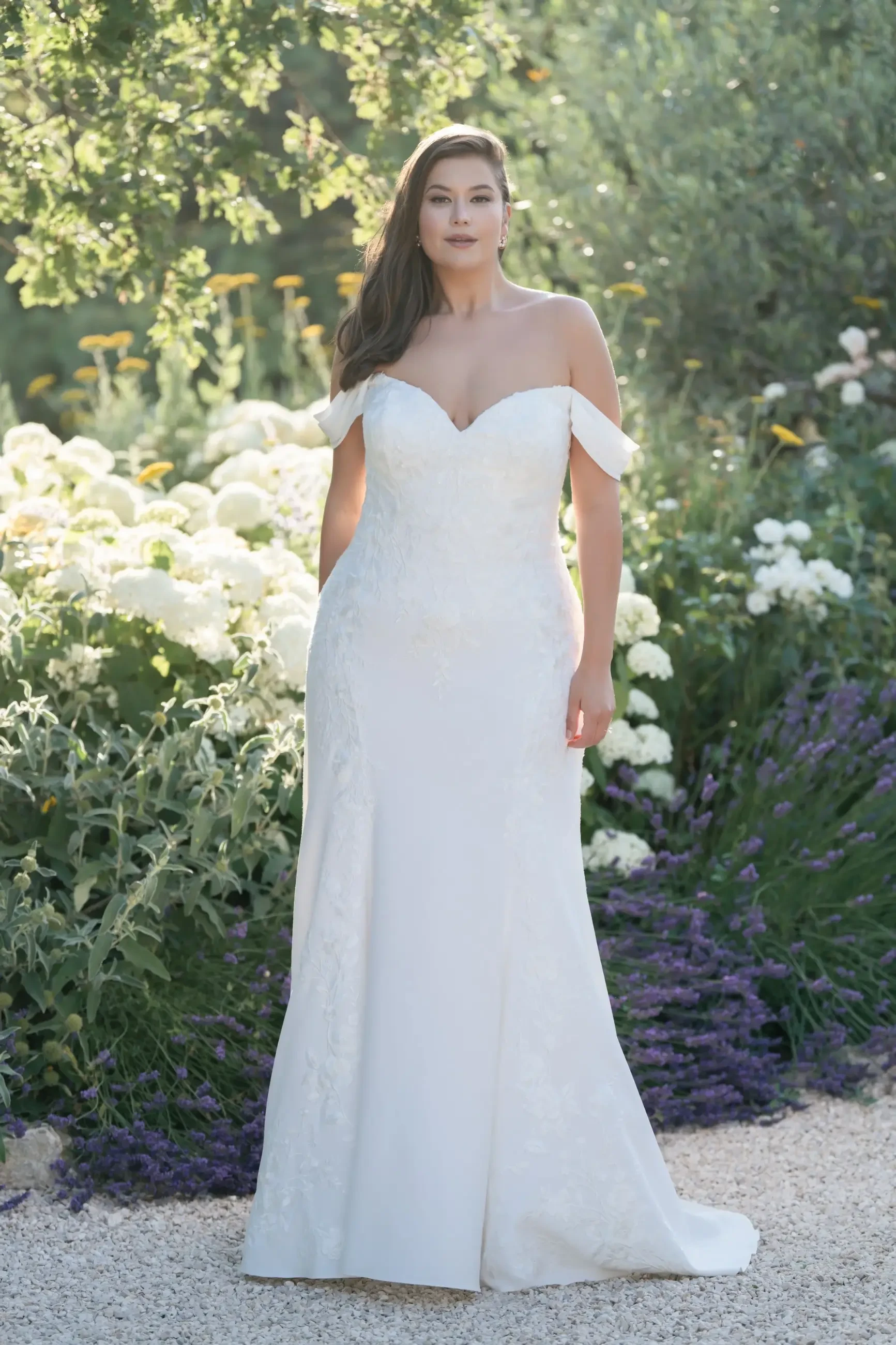 Model is wearing a plus size off the shoulder crepe sheath bridal gown with lace paneling by allure bridals